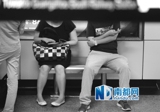 ↑在公园前站,两名乘客边等车边低头玩手机。